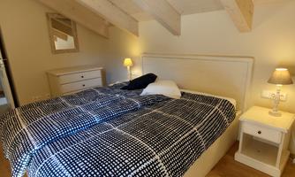 A cozy bedroom with a large bed and wooden beams on the ceiling. The decor is bright and modern, with a nightstand and a lamp.
