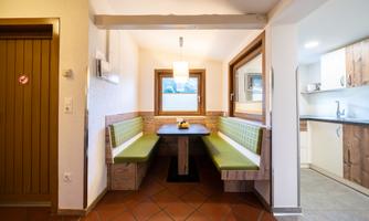 A cozy dining area with green benches and a wooden table. The bright kitchen is visible in the background.