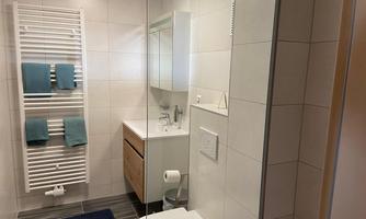 A modern bathroom with white tiles and a wooden vanity. Towels and a shower are also available.