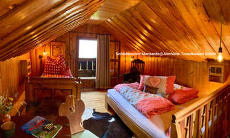 A cozy bedroom in the attic with wooden paneling. There is a bed with colorful pillows, a small table, and plenty of daylight.
