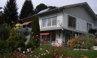 A beautiful house with a garden full of colorful flowers. In the background, there are trees and a terrace with a sunshade.