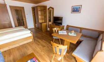 A cozy room with two beds, a dining table, and a seating area. The decor is made of wood, and the room has large windows.