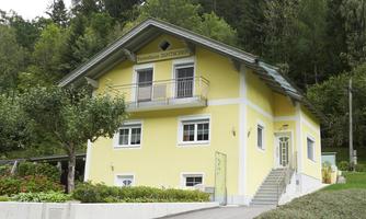A beautiful yellow house with a balcony, surrounded by trees. The path leads to the entrance of the house, which is situated in a tranquil landscape.