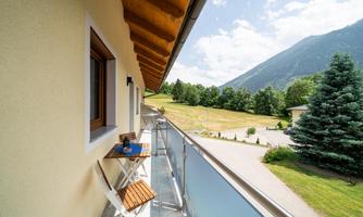A balcony with a small table and chairs, surrounded by a picturesque landscape. In the background, mountains and green trees can be seen.