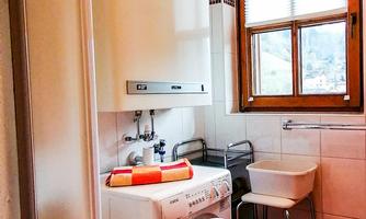 A small bathroom with a shower and a washing machine. There is a tidy kitchen with a window that lets in daylight.
