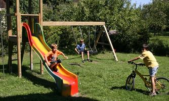Three children are playing in a playground. A boy is sliding down a slide, while the others are playing on a swing and a bicycle.