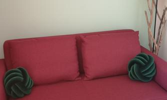 A red sofa with two green cushions is positioned in front of a light green wall. Next to the sofa, there is a lamp and a decorative branch.