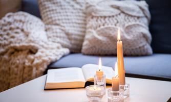 A cozy living space with an open book and burning candles on a table. In the background, soft cushions and a blanket can be seen.