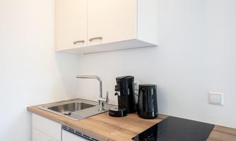 A modern kitchen with a sink, coffee machine, and stove. The furniture is in light colors and the countertop is made of wood.