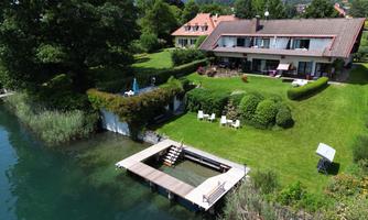 A large house with a well-maintained garden by the water. A wooden terrace with access to the water complements the idyllic setting.