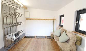 A modern room with wooden flooring and white walls. On the left are coat racks and on the right a cozy seating area with cushions.