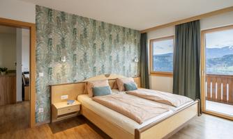 A modern bedroom with a double bed and elegant wood design. The walls are decorated with a plant pattern, and the large windows provide a beautiful view.
