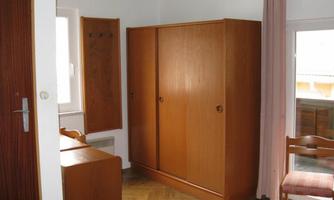 A simple room with wooden furniture. There is a wardrobe and a table next to a window.