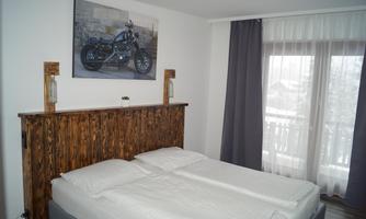 A cozy bedroom with a wooden headboard and a large double bed. An artwork featuring a motorcycle hangs on the wall, and large windows let in plenty of daylight.