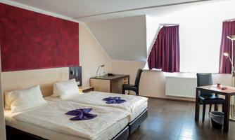 A cozy hotel room with two beds and a desk. The walls are painted in red and beige, and the room has large windows with red curtains.