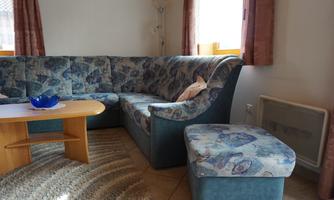 A cozy living room with a blue corner couch and a wooden table. The light shines through the window and the atmosphere is inviting.