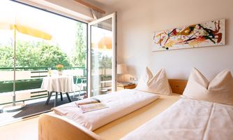 A modern bedroom with a double bed and two pillows. The balcony is equipped with a table and sun umbrellas, and there is plenty of daylight.