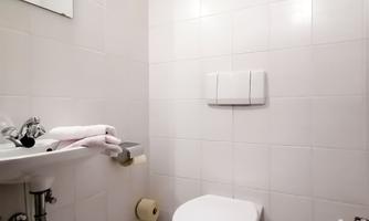 A small, white-tiled bathroom with a sink and a toilet. Clean towels are lying on the sink.