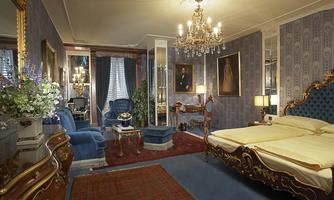 An elegant hotel room with blue walls and tasteful decor. The room is equipped with a chandelier, antiques, and a cozy seating area.