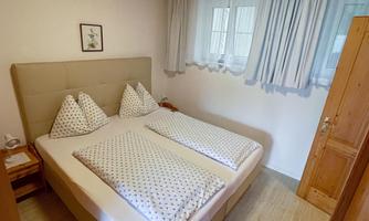 A cozy bedroom with a double bed and two pillows. The walls are light, and there are large windows with curtains.