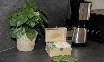 A small kitchen corner with a plant and a coffee pot. Next to the coffee pot is a wooden box with various tea bags.