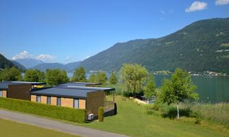 A peaceful landscape with modern wooden houses by the shore of a lake. In the background, green mountains rise under a bright blue sky.