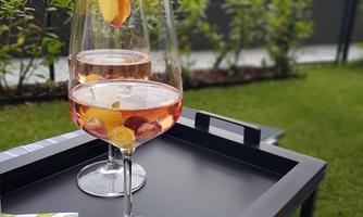 Two wine glasses with a pink beverage are on a tray in a green garden. In the background, bushes and a fence can be seen.