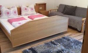 A cozy bedroom with a double bed and a gray sofa. The floor is fitted with a soft blue carpet.