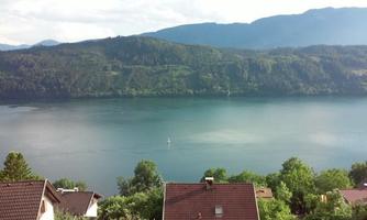 A quiet lake surrounded by green hills and trees. In the foreground, there are some houses with red roofs.