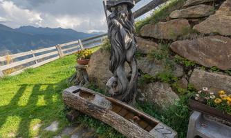 A beautiful wooden fountain with an artistic sculpture stands on a meadow. In the background, there are rocks and a wooden fence visible under a cloudy sky.