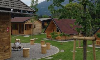A cozy park landscape with several wooden houses and wooden seating areas. In the background, green mountains and a cloudy sky can be seen.