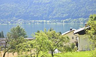 A picturesque landscape with a tranquil lake and gentle hills. In the foreground, there are trees and a charming house.