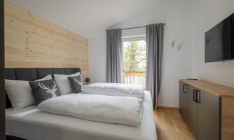 This cosy and modern bedroom exudes a pleasant sense of calm thanks to its light wood elements and subtle colours. The comfortable double bed with upholstered headboard and stylish decorative cushions with a deer motif invites you to relax. Floor-to-ceiling windows with a view of the countryside provide plenty of natural light and harmoniously connect the room with its surroundings. A simple wardrobe and a wall-mounted television complete the furnishings and offer additional comfort.’