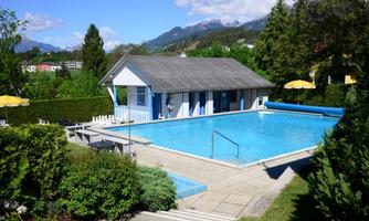 A beautiful blue pool in a garden with green trees. In the background, mountains and a light blue sky can be seen.