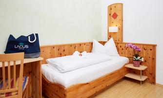 A cozy room with a single bed and wooden paneling. A bag and a potted plant are on the table, while fresh bed linen is ready.