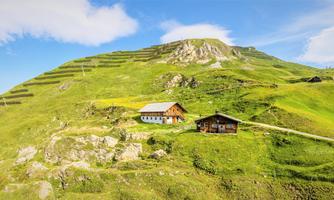 A picturesque hilly landscape with green meadows and two traditional Alpine houses. In the background, there is a clear sky and gentle mountains.
