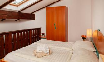 A cozy bedroom with a double bed and a large window. The room is bright and features wooden details as well as a dresser.