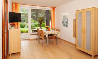 A bright dining room with a table and chairs, surrounded by windows. In the background, there is a garden with trees.