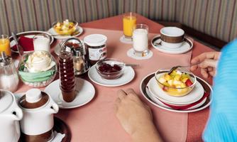 A set table with a selection of breakfast dishes, including fruit salad and drinks. A person is reaching for a bowl of fresh fruit.