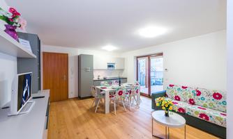 A modern living room with a dining table and a colorful sofa. The room has wooden flooring and large windows that let in plenty of light.
