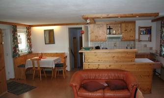 A cozy living space with a brown leather couch and a wooden table. The kitchen is open and bright, with an inviting atmosphere.