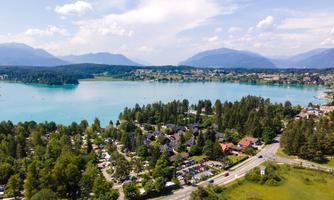 A picturesque landscape with a clear, blue lake surrounded by green trees. In the background, gentle mountains and a bright blue sky are visible.