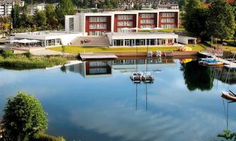 A modern building on the shore of a tranquil lake. In the background, hills and trees can be seen.