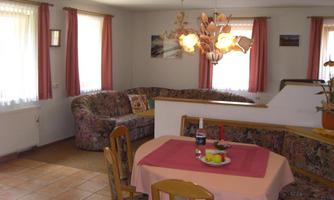 A cozy dining area with a round table, chairs, and fresh fruits. In the background, there is an inviting living room with a comfortable sofa and natural light sources.