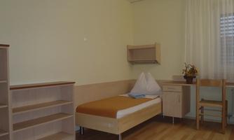 A simple room with a single bed, a table, and a chair.  
On the wall, there is a shelf and windows with curtains.