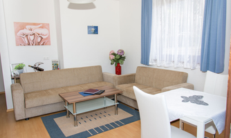 A bright living room with two beige sofas and a table in the middle. Pictures hang on the wall, and there is a dining table with four chairs.