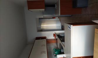 A modern kitchen with light wood and white cabinets. There is a table and access to another room.
