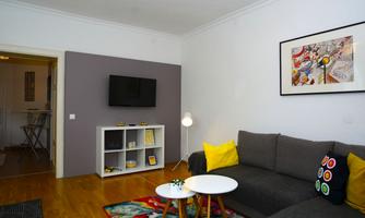 A modern living room with a gray sofa and colorful cushions. On the wall, there is a television and a decorative table with a small plant.