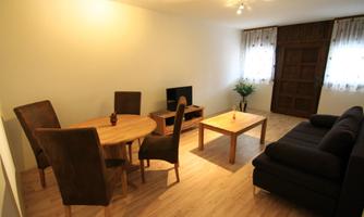 A cozy living room with a sofa, a dining table, and chairs. Plenty of light shines through the windows and the floor is made of wood.