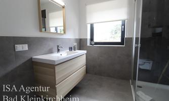 A modern bathroom with a wood-colored washing machine and a large shower. The room is bright and friendly with a window that lets in daylight.